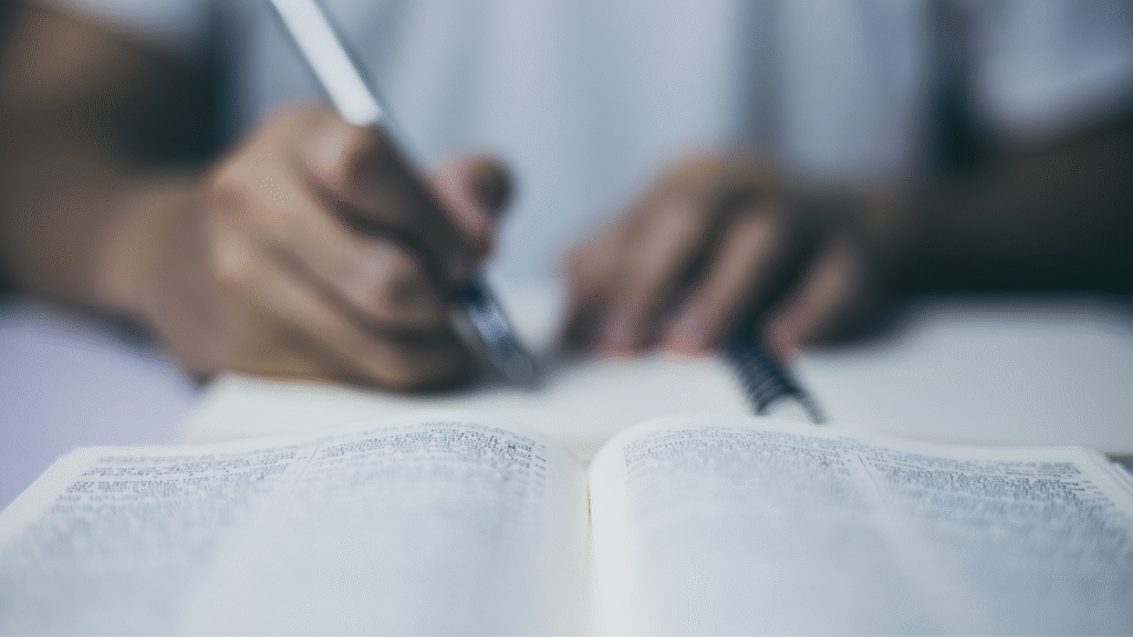 Hands holding a pen in front of a book and notebook.