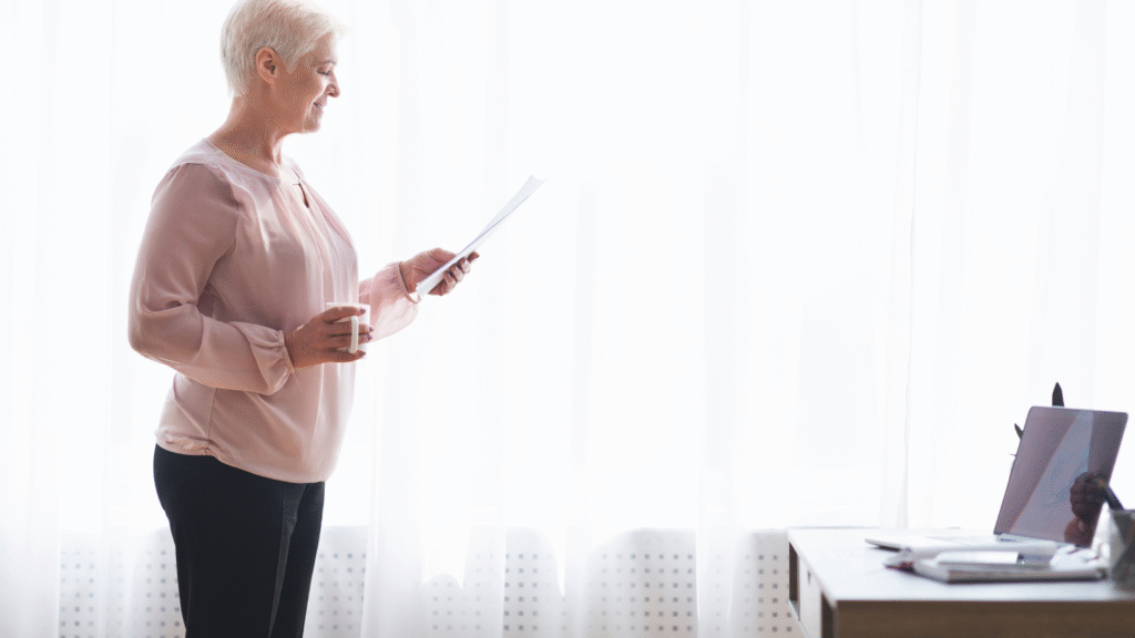 An old woman in a pink blouse smiling at a paper she is holding while also holding a cup of coffee.